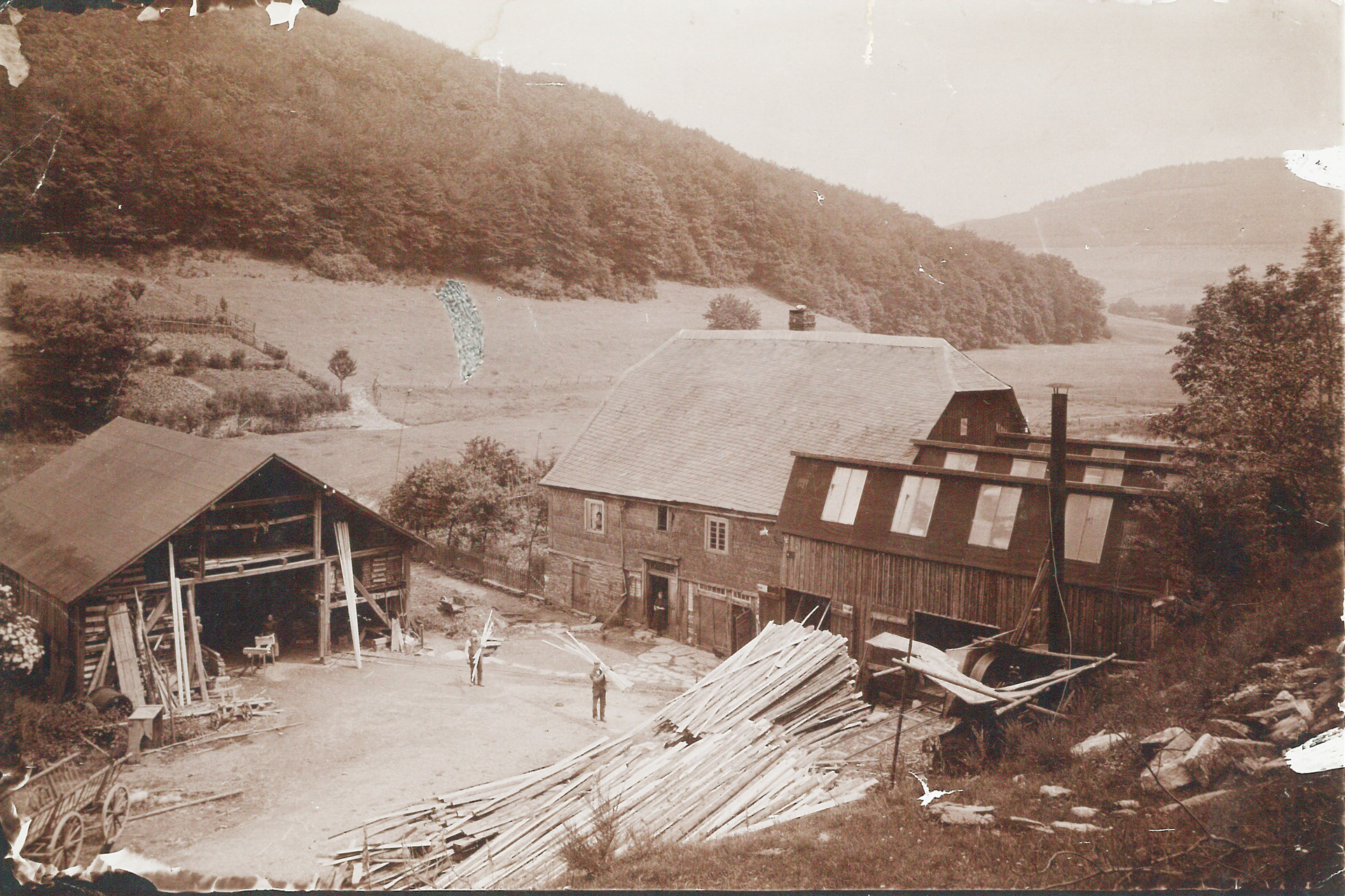 Altes Sepia-Foto der Mühle auf Gut Hanxleden mit angrenzender Scheune, Holzstapeln und zwei Arbeitern im Hof, umgeben von bewaldeten Hügeln.