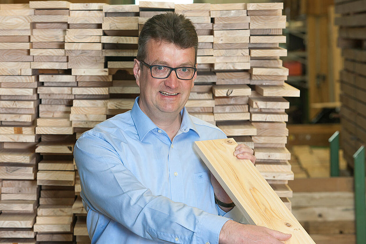 Mann in blauem Hemd, hält eine Holzdiele in der Hand; im Hintergrund gestapelte Holzbohlen